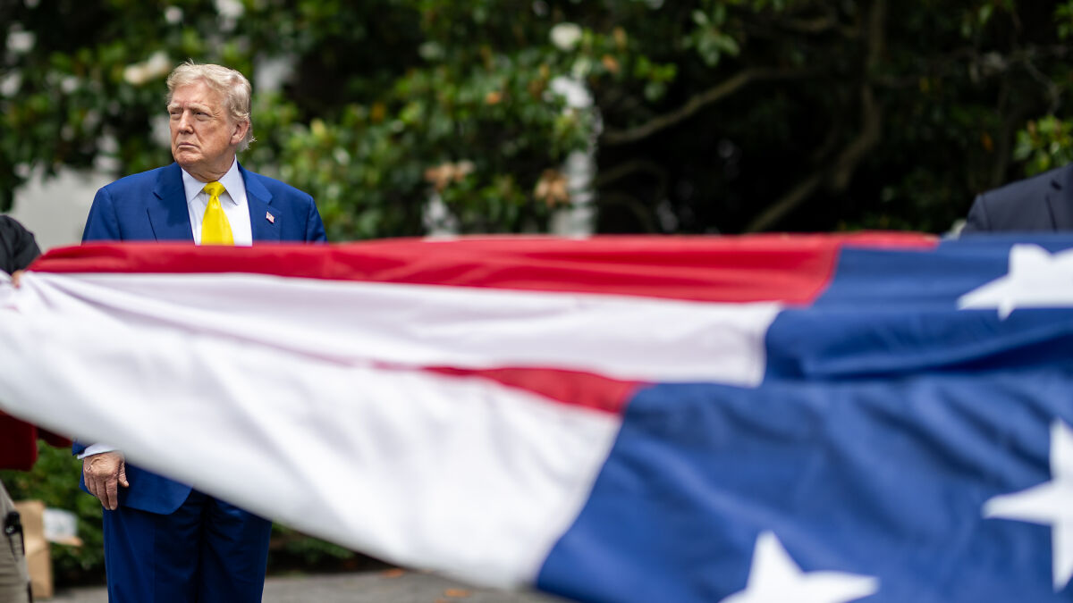 Former President Trump standing near a large American flag outdoors with trees in the background