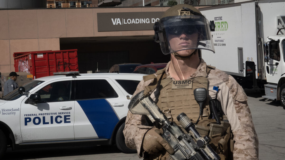 USMC soldier in tactical gear stands near Homeland Security police vehicle at parking lot entrance near stadium.