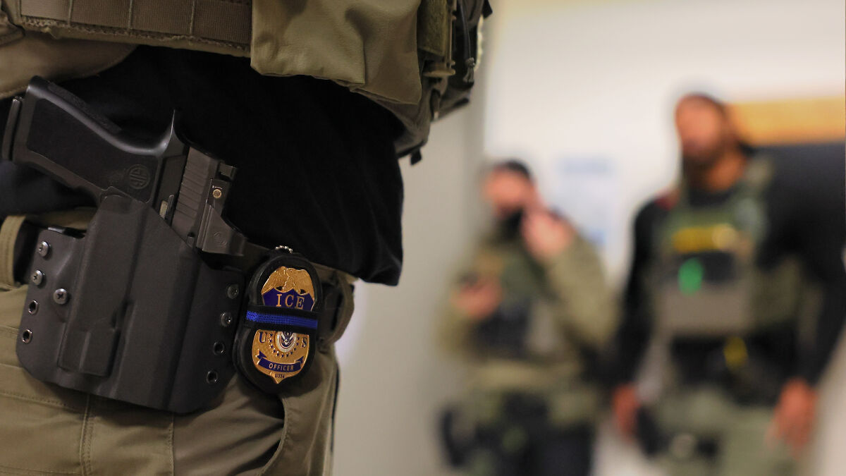 ICE officer with badge and holstered gun in focus, agents blurred in background during enforcement operation.