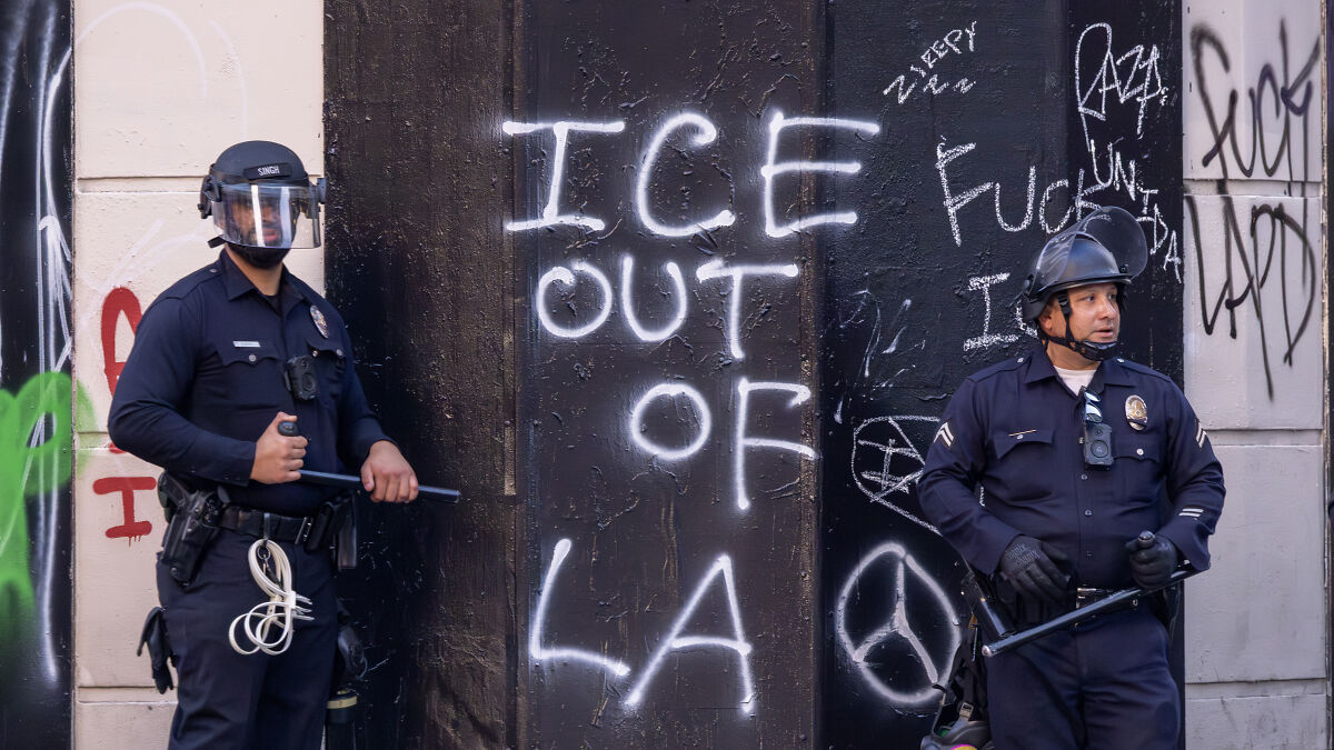 Two police officers in riot gear stand by a wall with graffiti including ICE out of LA amid California gang tensions.