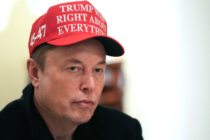 Elon Musk wearing a red political cap, looking thoughtful in a close-up image related to Musk and Trump rivalry and friendship.