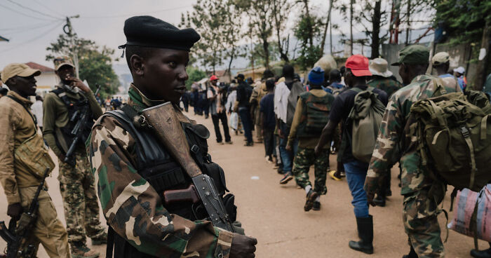 Armed soldier standing guard as a long line of people wait, illustrating deadly wars and humanitarian crises in conflict hotspots.