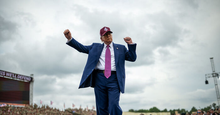 Donald Trump wearing a suit and hat, raising arms at an outdoor event, with cloudy sky in the background.