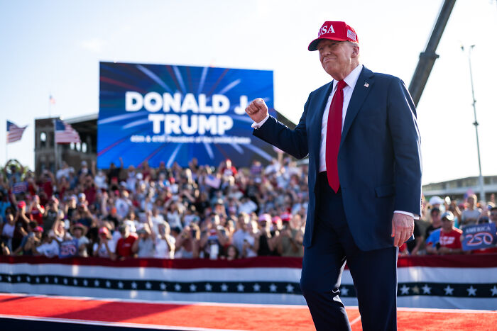Donald Trump wearing a red USA hat giving a fist pump at a crowded outdoor rally with a large screen in the background.