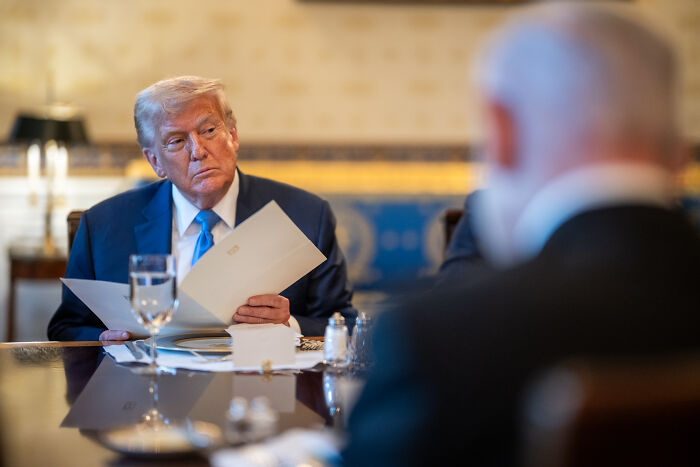 Donald Trump sitting at a table holding documents, reacting during a discussion as MAGA supporters react to new Epstein findings.