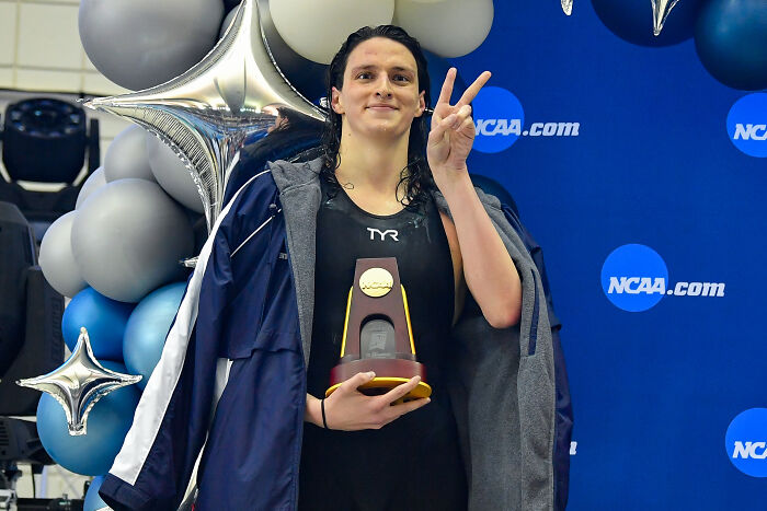 Trans swimmer Lia Thomas holding a trophy and making a peace sign at an NCAA swimming event with balloons behind.