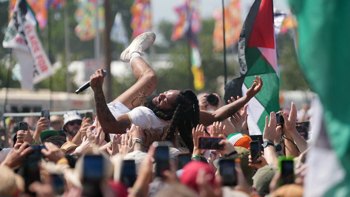 Crowd surfing by rap artist Bob Vylan during a live outdoor performance with fans holding up phones and flags.