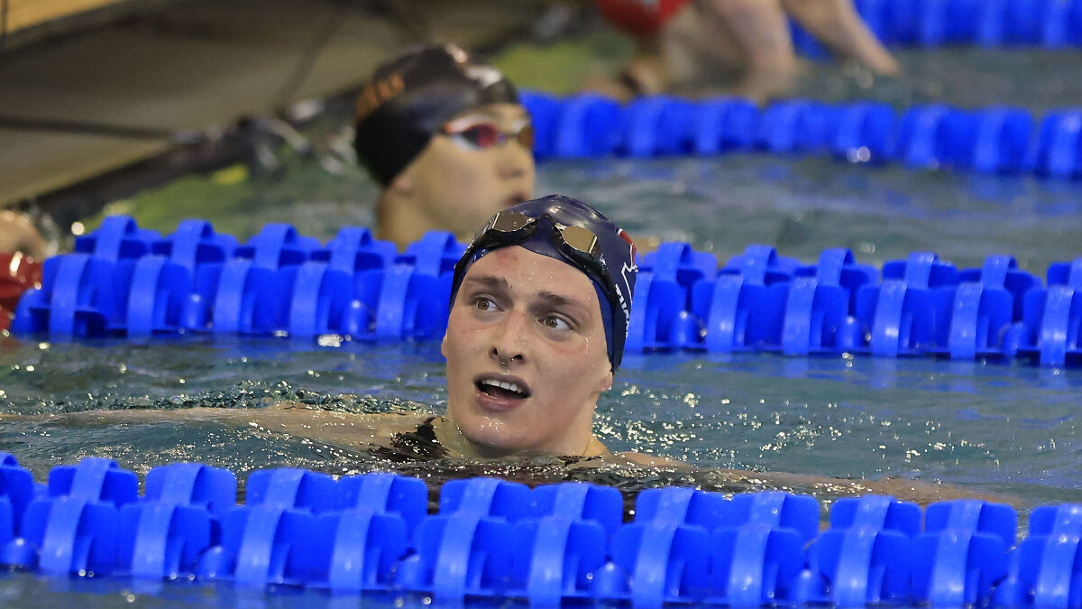 Trans swimmer Lia Thomas in swimming pool during competition, wearing swim cap and goggles, looking to the side.