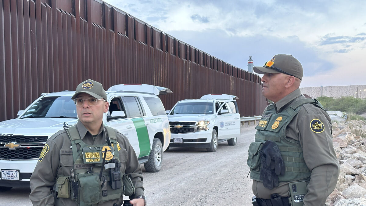 Two U.S. Border Patrol agents near vehicles by the border fence at the U.S.-Mexico border addressing asylum issues.