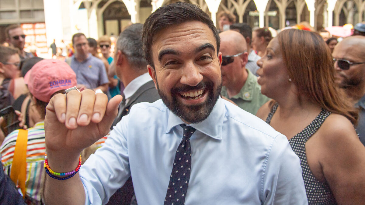 Zohran Mamdani smiling in a crowd, wearing a white shirt and polka dot tie, during Columbia application controversy event.