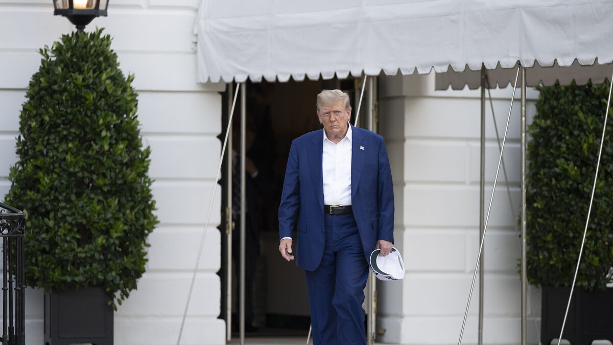 Donald Trump walking outside the White House, holding a hat in hand with a serious expression on his face.