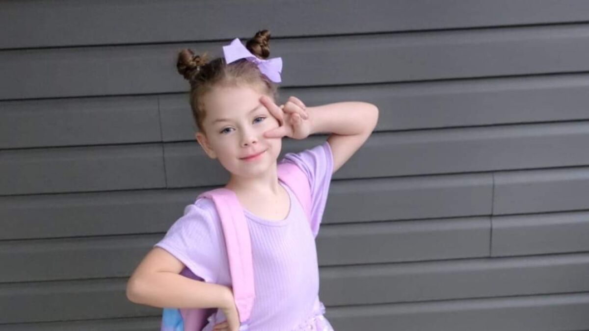 Young girl posing with peace sign, wearing purple outfit and backpack, related to drowning in crowded swimming pool incident.