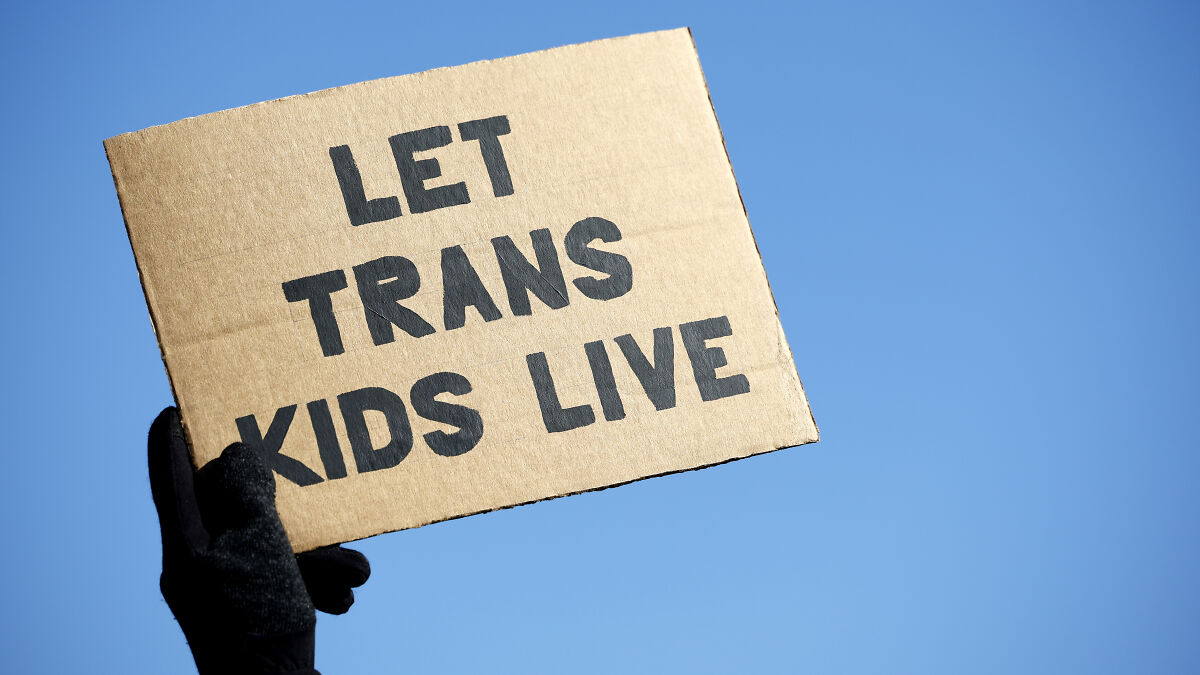 Protest sign reading let trans kids live held against clear blue sky, related to doctors providing trans health care debate.